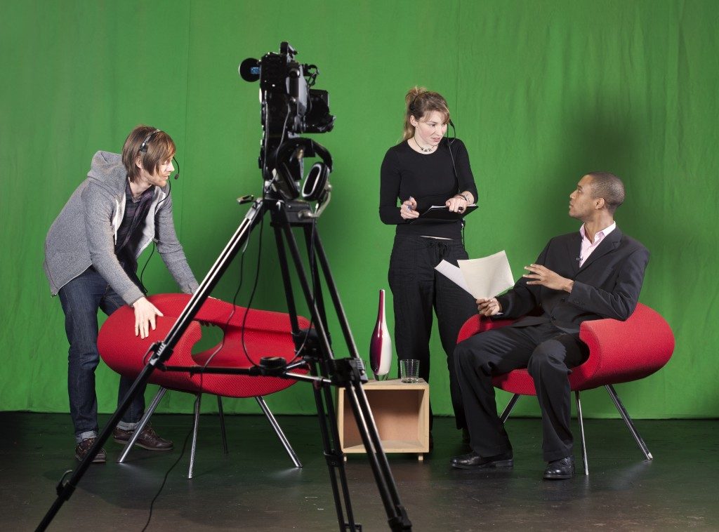 A crew member arranges furniture whilst the floor manager talks to the presenter on a TV studio set