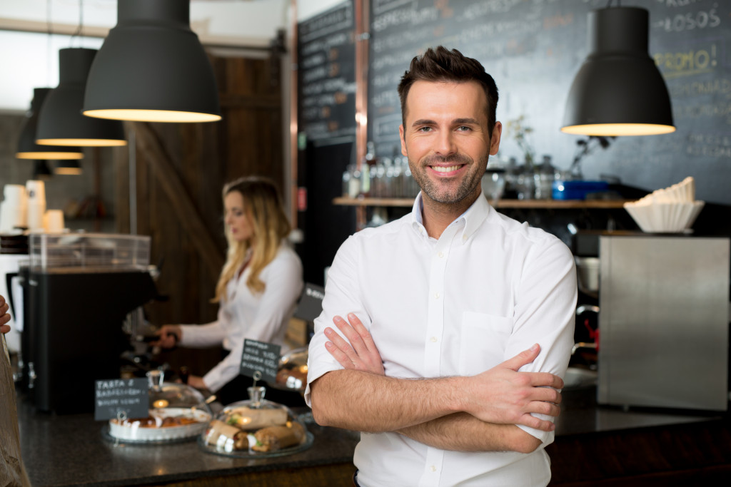 Successful small business owner standing with crossed arms with employee in background preparing coffee