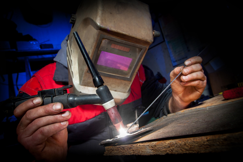 A close-up of a welder wearing a welding mask and using a TIG gun to weld metal to a surface