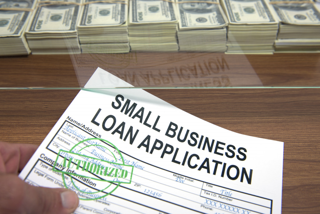 A close-up of a hand and an approved small business loan application in front of a glass window with stacks of dollars behind it