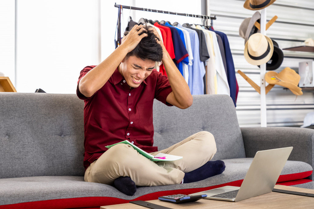 A business owner sitting cross-legged on a couch clutching his head in frustration while a laptop, calculator, and a ledger are in front of him