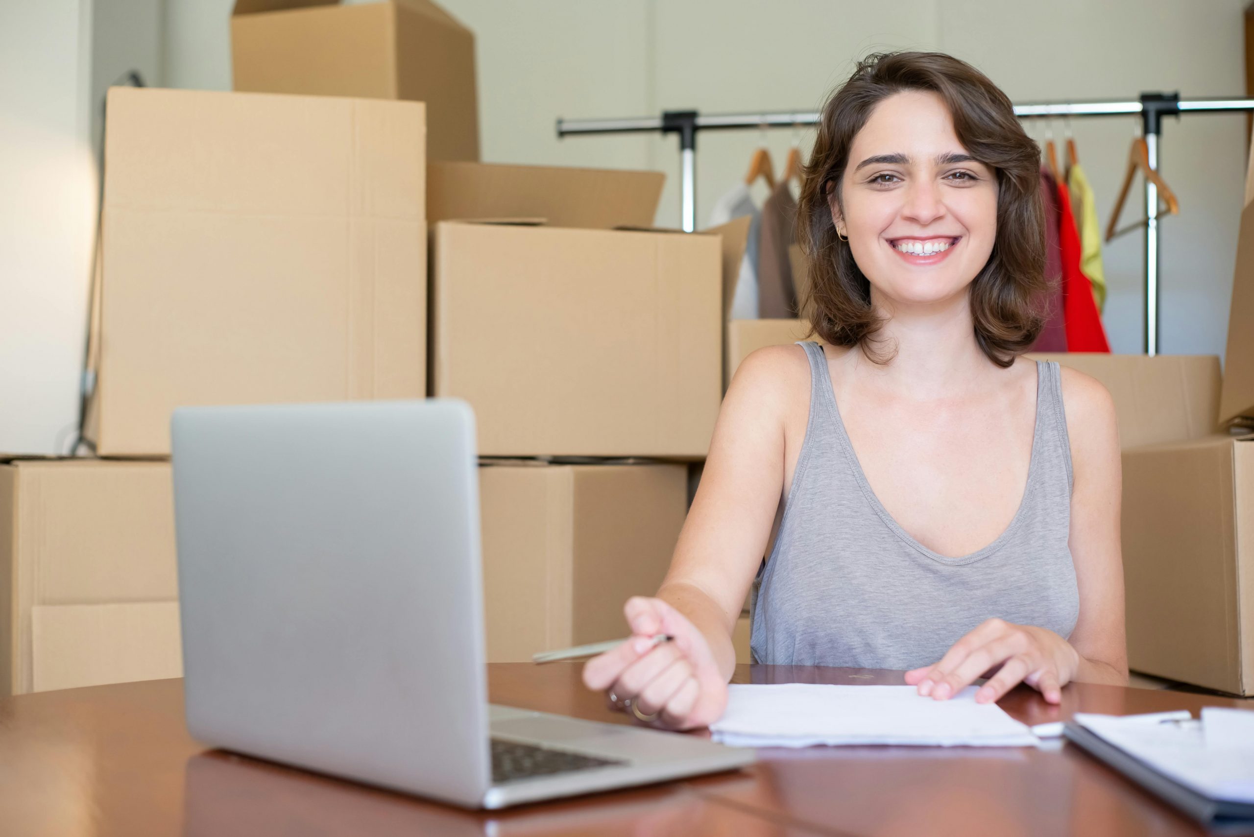 entrepreneur smiling at her desk, with a laptop in front of her and shipping boxes on shelves behind her